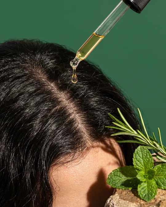 A dropper of oil at the part of dark hair, with rosemary and mint