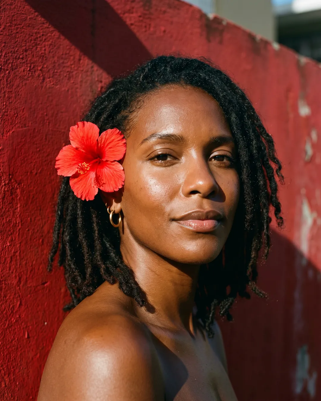 A woman with locs and a red flower behind her ear against a red wall